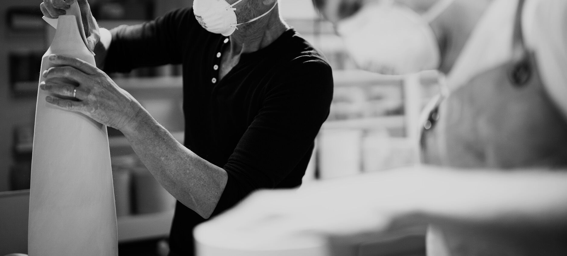 Black-and-white photo of a masked craftsperson shaping a large ceramic lamp in a studio while another person works in the foreground.