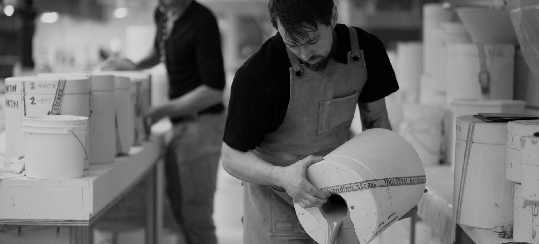 Black-and-white photo of a craftsman pouring ceramic slip into a large lamp mold inside a working pottery studio.